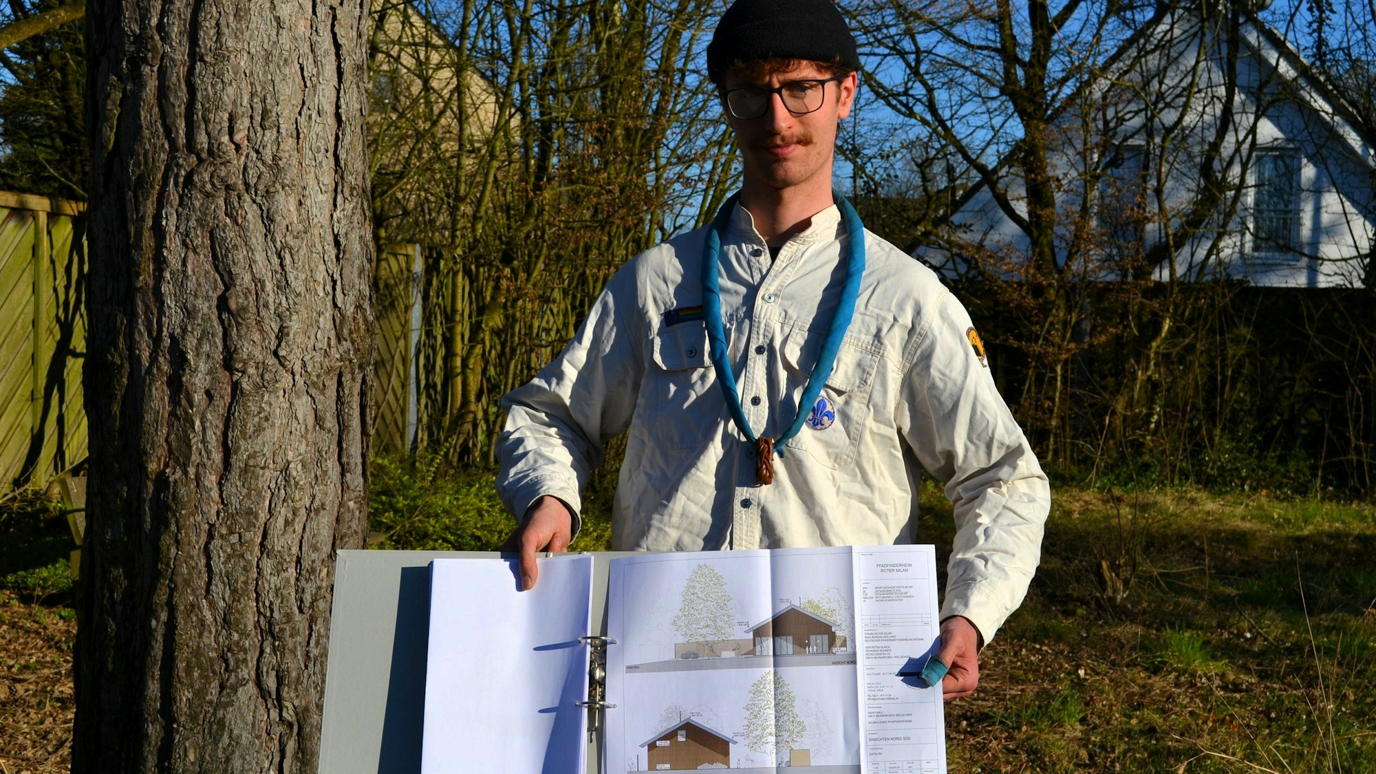 Ein junger Mann in Pfadfinderhemd zeigt Planzeichnungen für ein Gebäude.