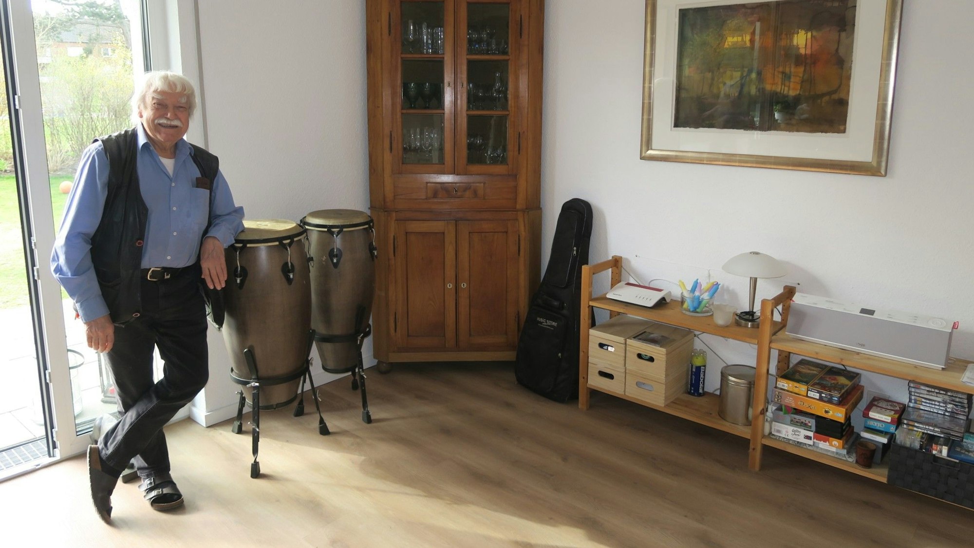 Ein älterer Mann mit weißem Haar und Schnurrbart steht in einem hellen Raum auf zwei Bongo-Trommeln gelehnt.