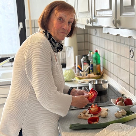 Eine Frau mit kinnlangem Haar, die Pullover und Schal trägt, steht in einer Küche und schneidet eine Paprika.
