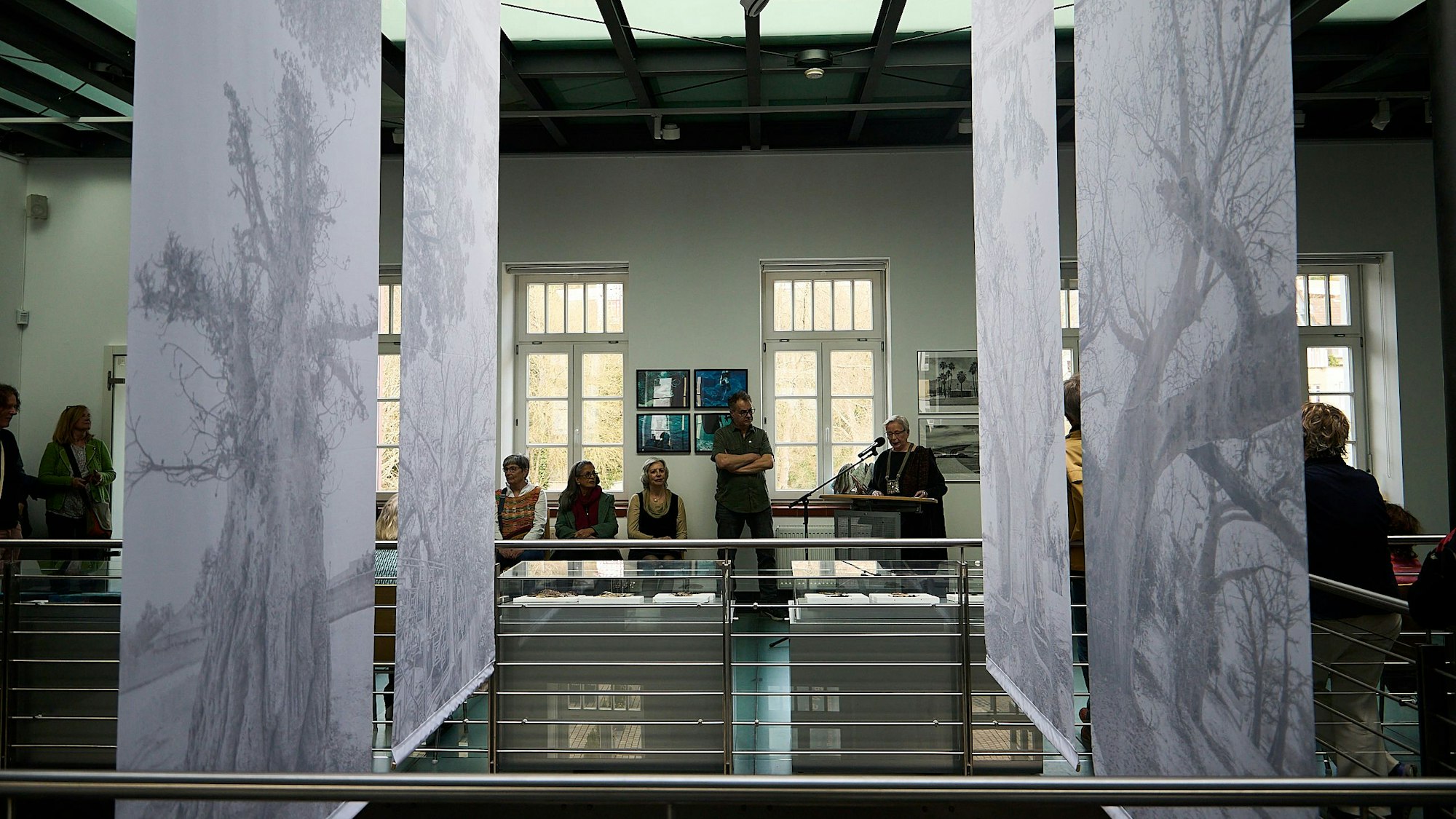 Fotografien von Rendel Freude hängen in der Mitte des Kunstforums. Im hinteren Bereich sind Menschen zu sehen. Eva-Maria Hermanns steht an einem Pult und spricht in ein Mikrofon.