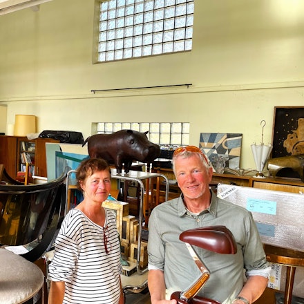 Eine Frau im Ringelshirt und ein Mann mit grauem Haar stehen in einem Lagerraum voller Möbel und Deko-Objekte.