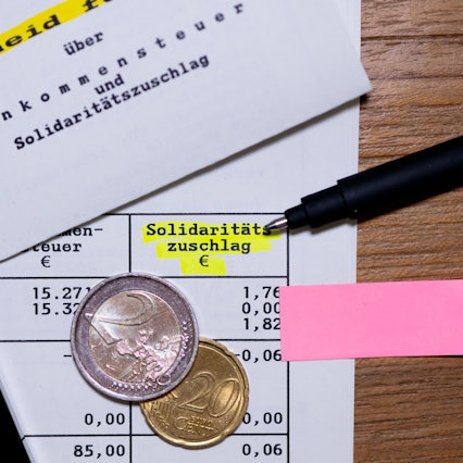 Das Wort „Solidaritätszuschlag“ ist neben einem Taschenrechner und einem Stift auf einem Steuerbescheid zu sehen. (Symbolbild)