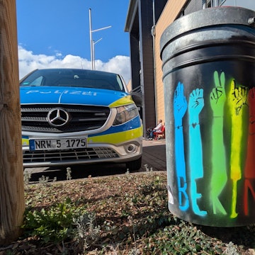 Der englischsprachige Schriftzug „Be kind“ ist auf einer in verschiedenen Farben besprühten Mülltonne zu lesen. Links steht ein Einsatzfahrzeug der Polizei.