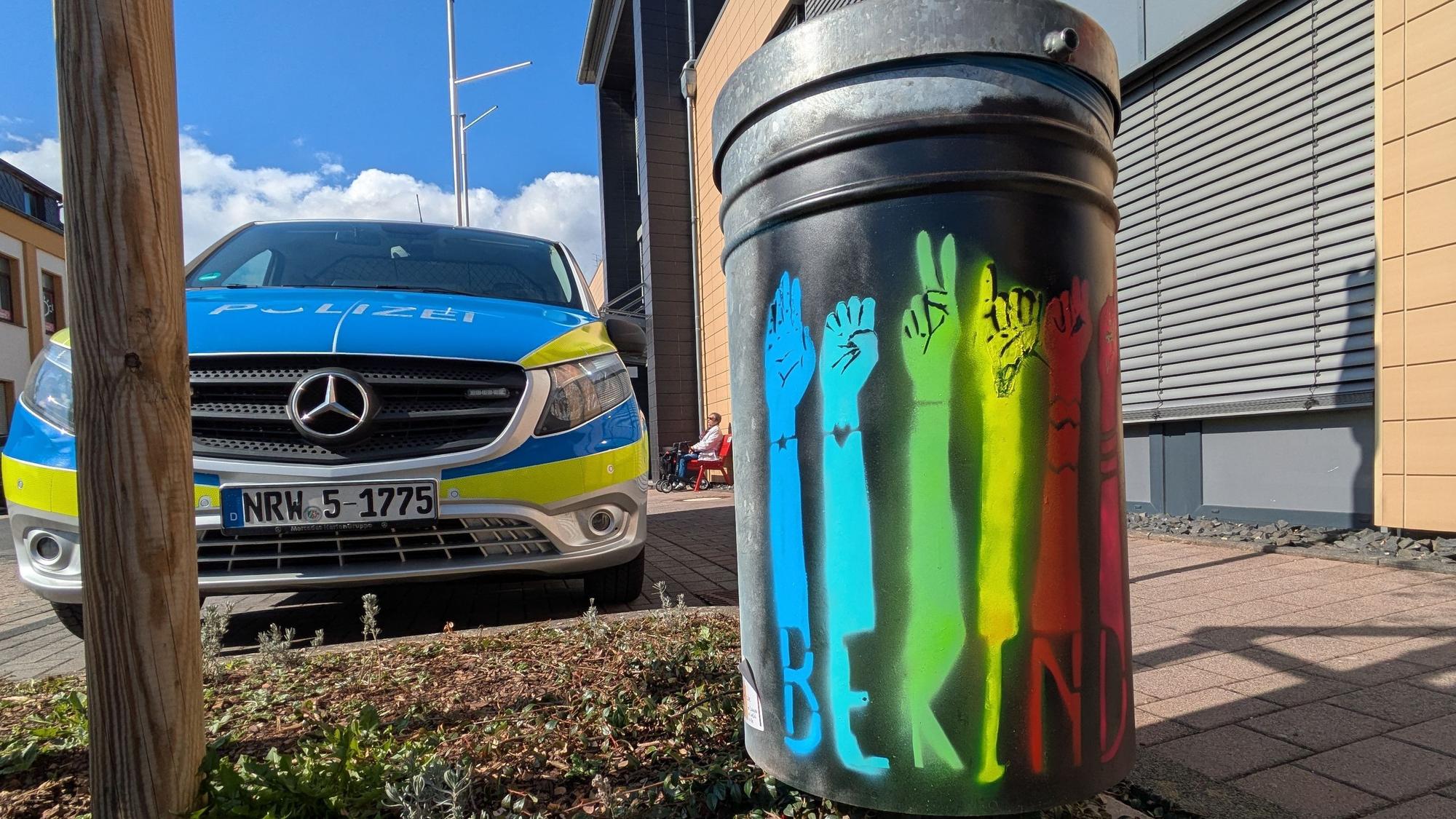 Der englischsprachige Schriftzug „Be kind“ ist auf einer in verschiedenen Farben besprühten Mülltonne zu lesen. Links steht ein Einsatzfahrzeug der Polizei.