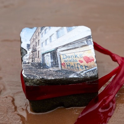 Das Symbolbild zeigt einen Pflasterstein, den Jochim Rieger in Bad Münstereifel mit einem Foto gestaltet hat. Es zeigt ein Fenster nach der Flutkatastrophe 2021, das mit Brettern verschlossen ist und auf dem mit Herzchen ein Dank an die Helfer zu lesen ist.