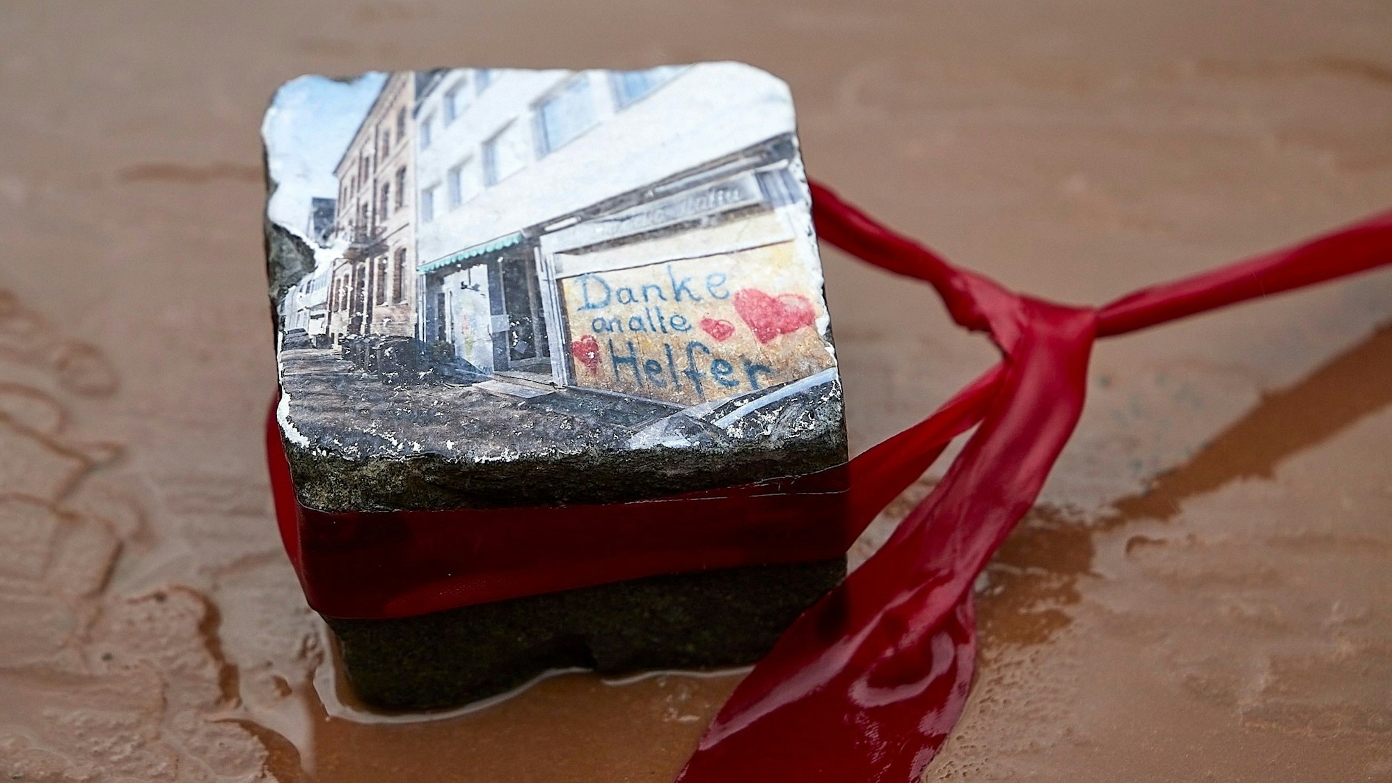 Das Symbolbild zeigt einen Pflasterstein, den Jochim Rieger in Bad Münstereifel mit einem Foto gestaltet hat. Es zeigt ein Fenster nach der Flutkatastrophe 2021, das mit Brettern verschlossen ist und auf dem mit Herzchen ein Dank an die Helfer zu lesen ist.