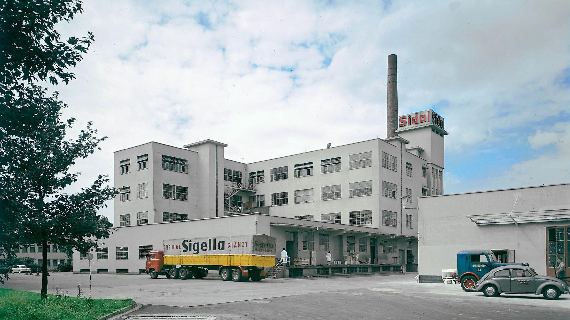 Die Sidol-Fabrik in Braunsfeld in den 1960er Jahren, ein weißes Fabrikgebäude mit einem Lkw davor.
