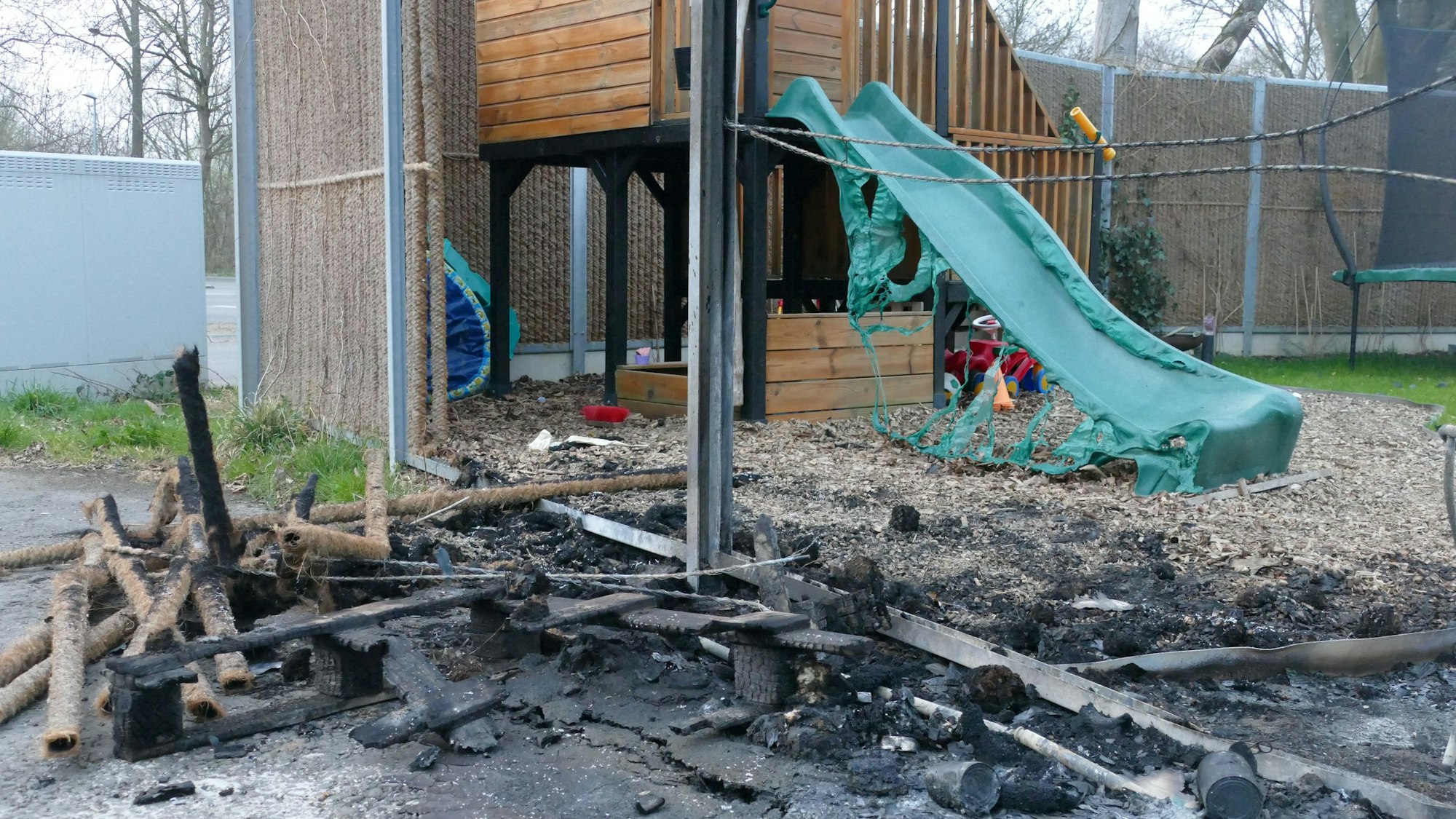 Brandspuren an einem Zaun, dahinter eine bizarr geschmolzene Kinderrutsche.