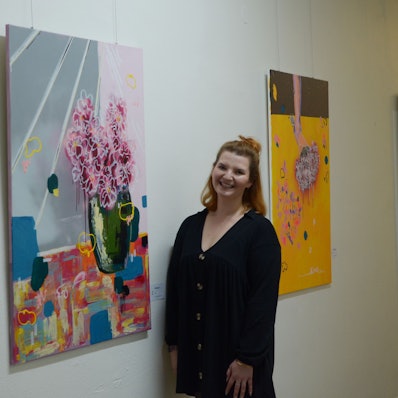 Das Bild zeigt Denise Wagner vor zwei Gemälden, die in einem Flur des Rathauses hängen.