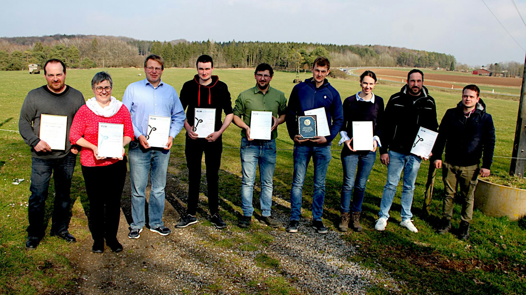 Neun Personen stehen in einer Reihe vor einer Wiese. Sie halten Urkunden in den Händen.