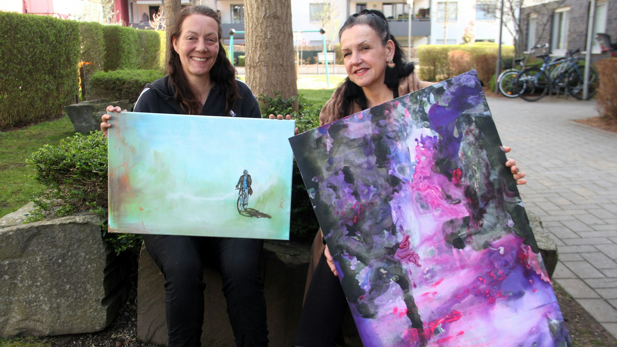 Zwei Frauen zeigen ihre Gemälde. Sie sitzen auf einem Baumbeet.