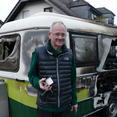 Ein Mann steht vor einem von Feuer völlig zestörten Wohnwagen.