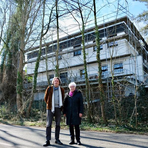 Ein Mann und eine Frau stehen vor einer Hangkante und einem Neubau. Vor dem Neubau stehen Bäume, die gefällt werden sollten.