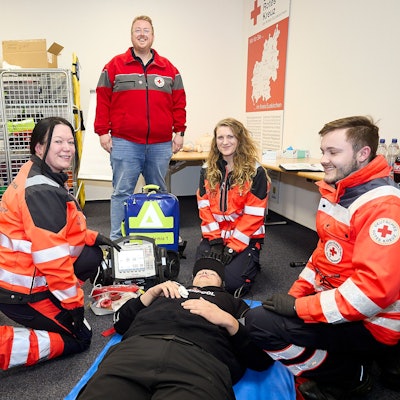 Zwei Frauen und ein Mann in Rettungsdienstkleidung knien im Rahmen ihrer Ausbildung in einem Schulungsraum vor einer Person. Der Akademieleiter steht und beobachtet sie.