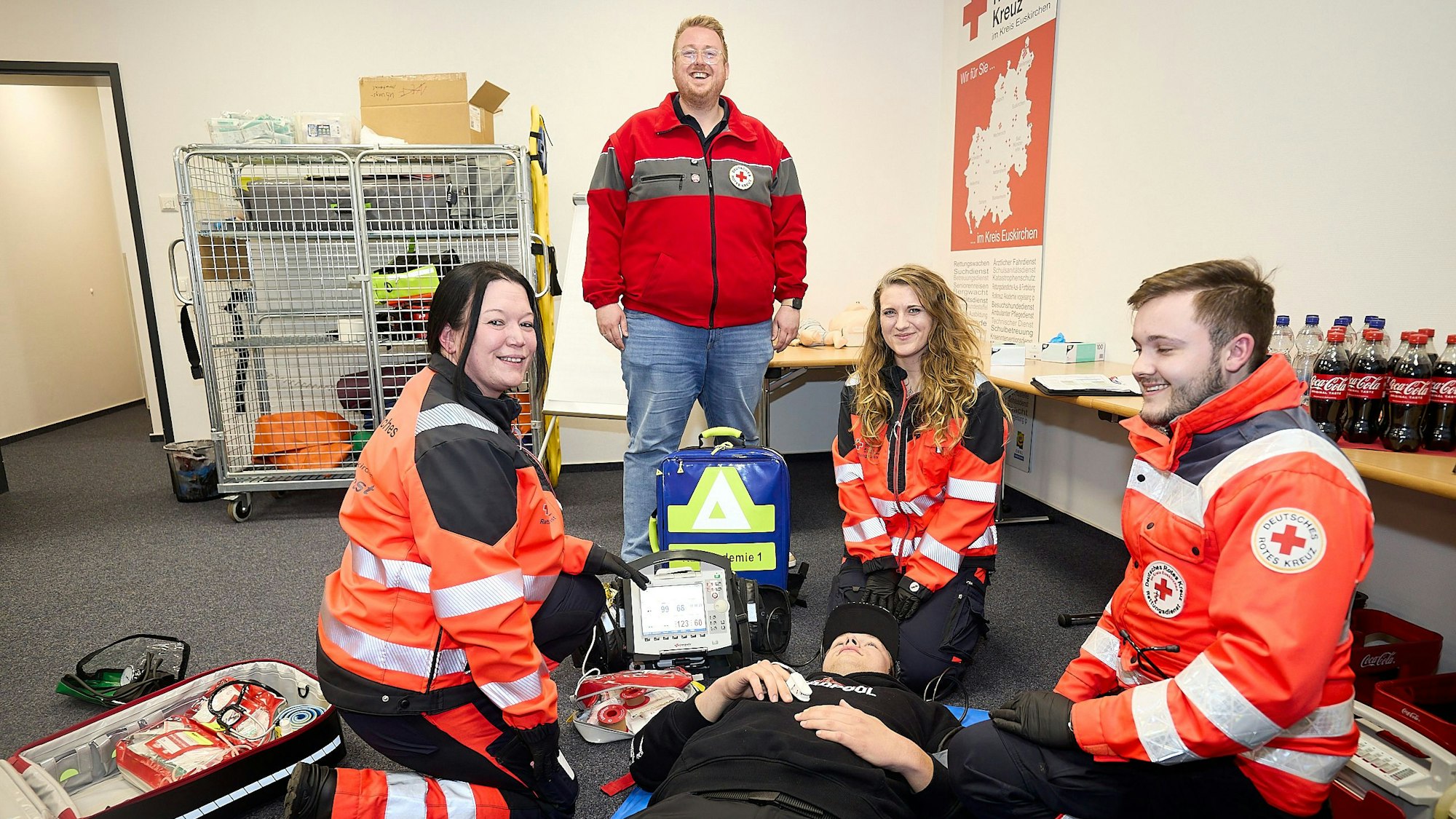 Zwei Frauen und ein Mann in Rettungsdienstkleidung knien im Rahmen ihrer Ausbildung in einem Schulungsraum vor einer Person. Der Akademieleiter steht und beobachtet sie.