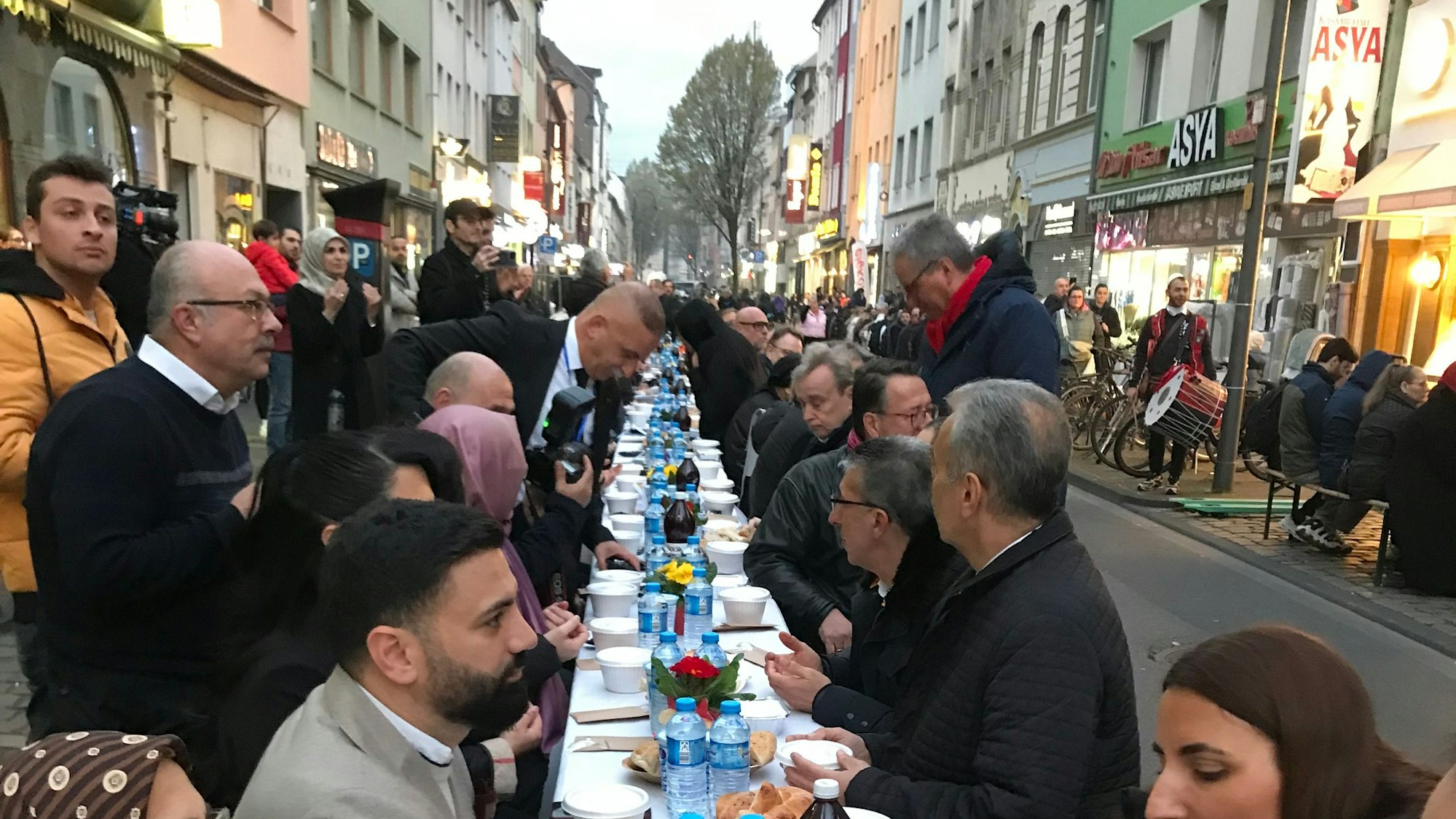 Viele Menschen an einem langen Tisch. Im Hintergrund die Geschäfte und Läden der Keupstraße.