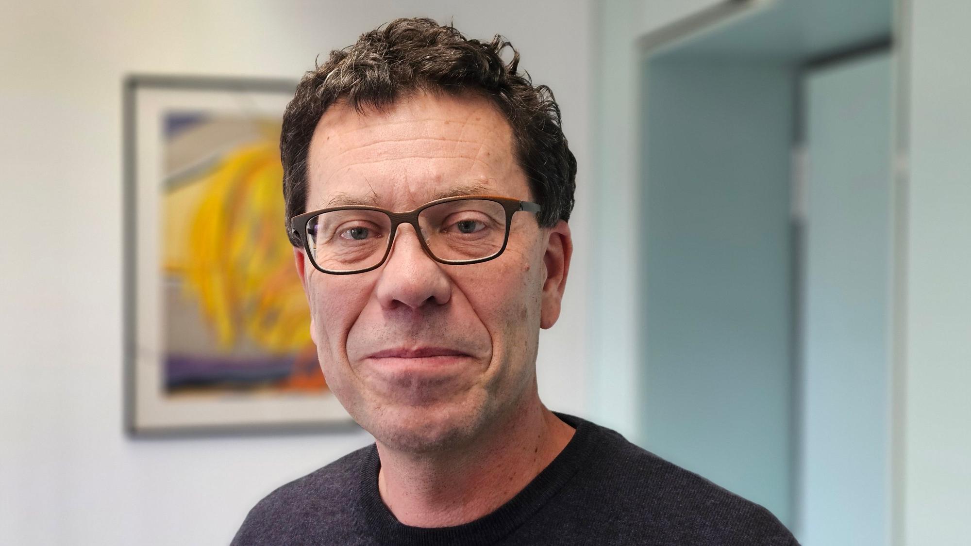 Auf dem Foto ist ein Mann mit Brille zu sehen. Er trägt einen dunklen Pullover.