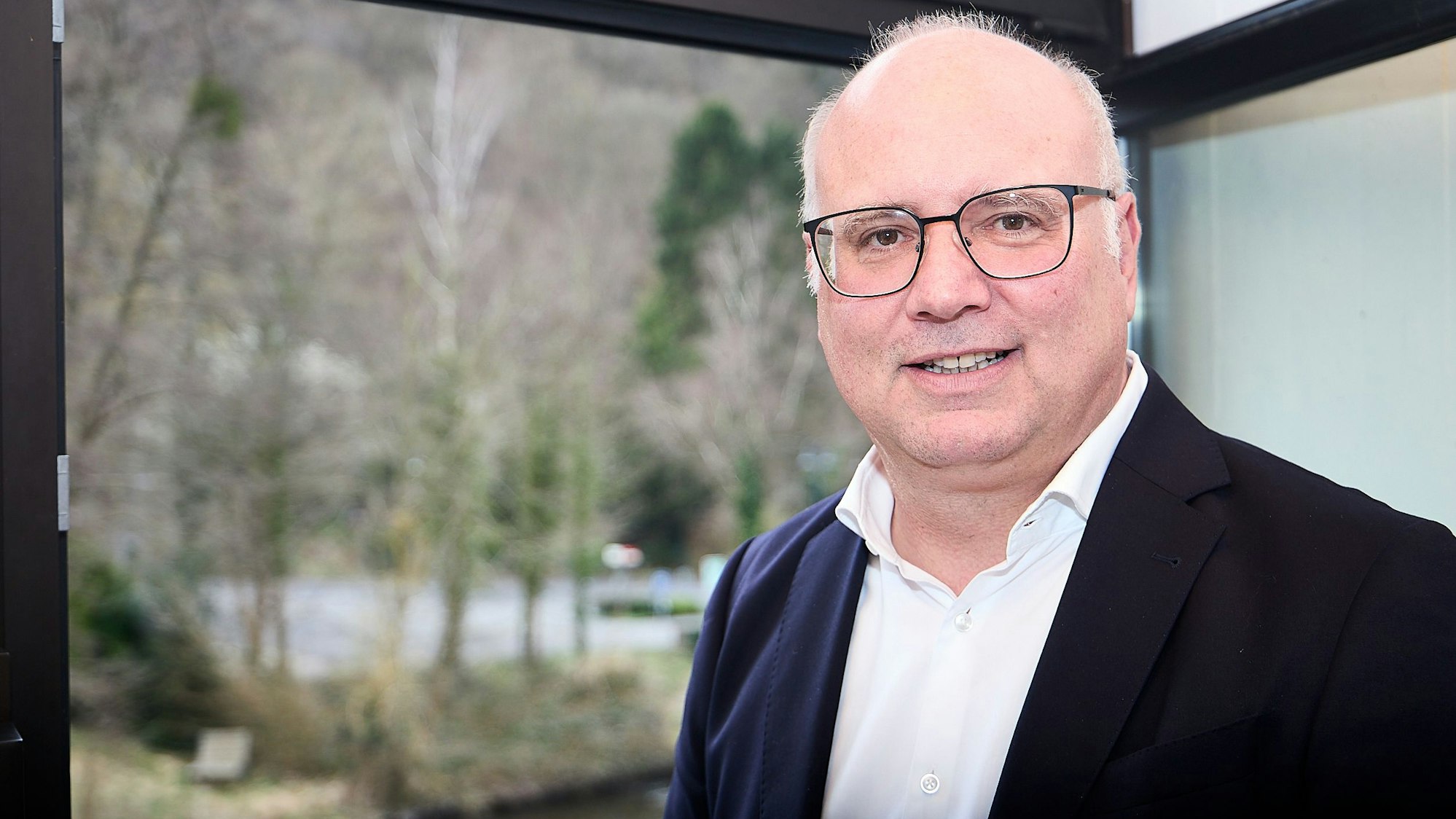 Ein Porträt des Bürgermeisters. Er steht vor einem Fenster mit Blick ins Grüne.