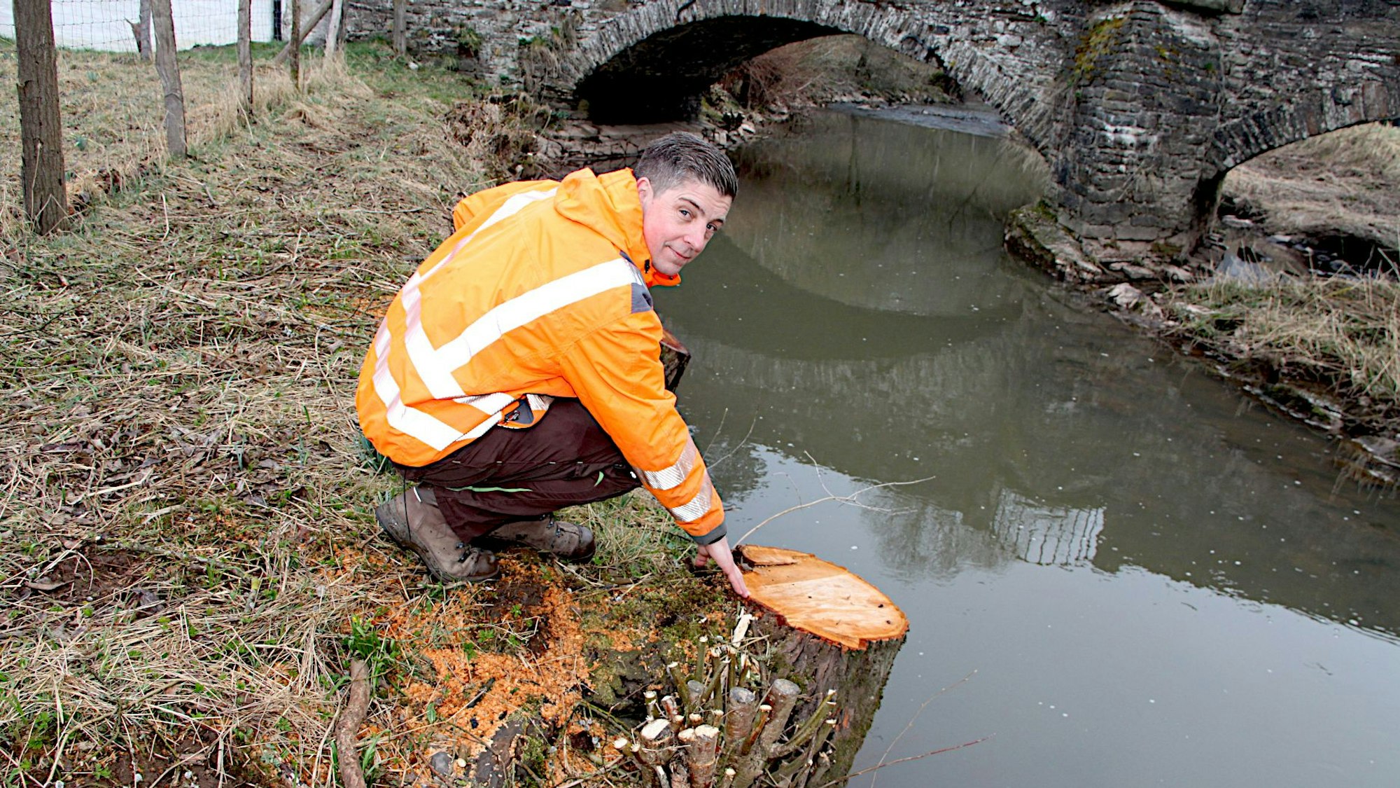 Der Baumkontrolleur Marco Dalboth hockt am Ufer der Kyll an einem Baumstumpf.