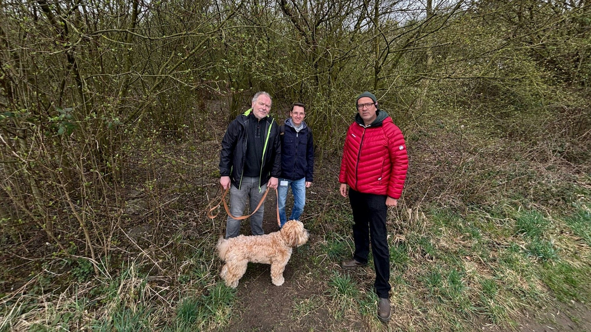 Auf dem Bild sind drei Männer und ein Hund zu sehen, die vor einem verwilderten Grundstück stehen.