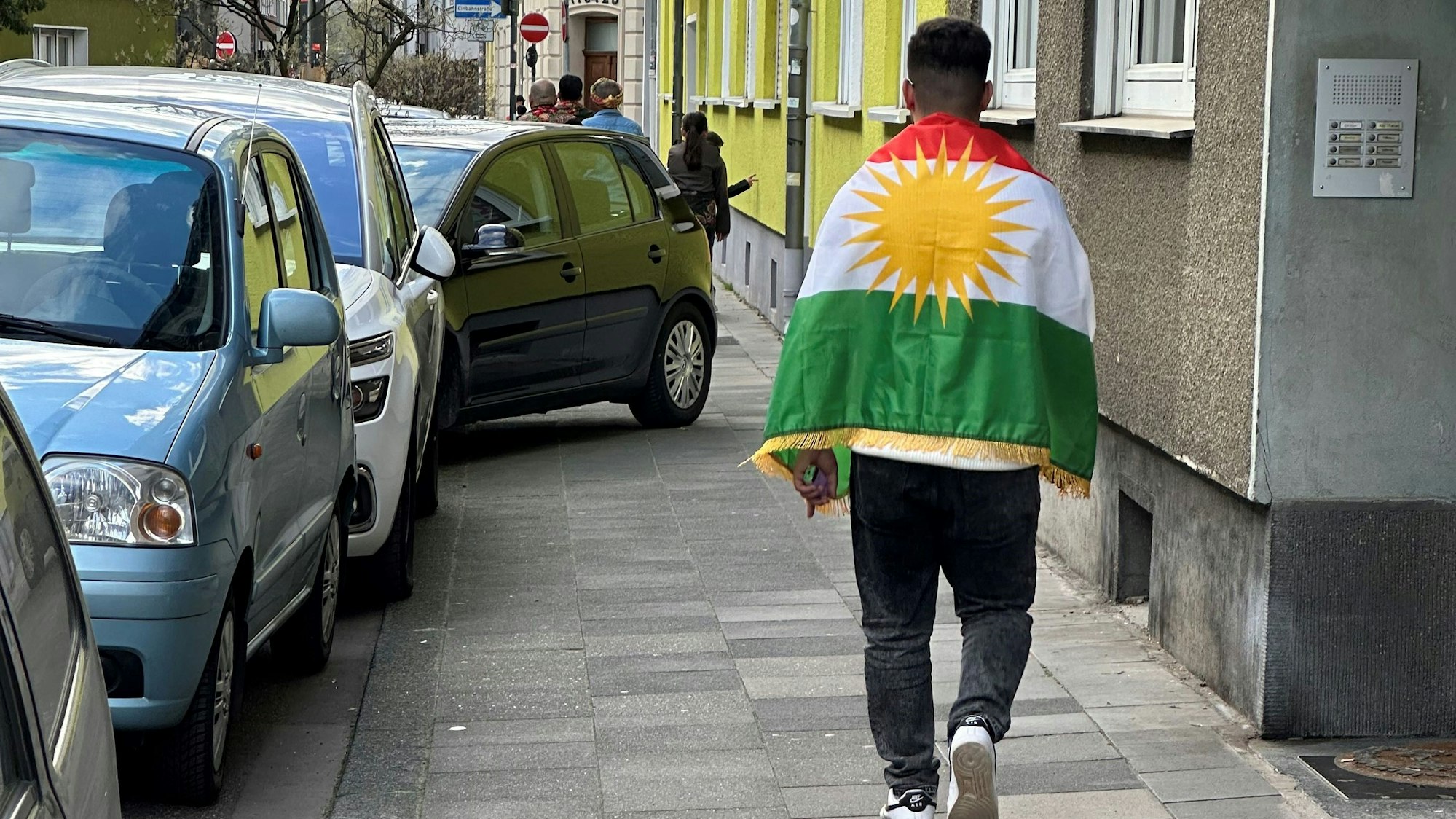 Ein mit einer kurdischen Fahne behangener Mann spaziert im Zuge des kurdischen Neujahrsfests durch Deutz, ein Auto im Hintergrund parkt halb auf dem Gehweg.