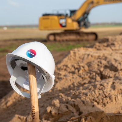 Ein Bauhelm ist auf einem Schaufelstiel abgelegt, im Hintergrund steht ein Bagger.