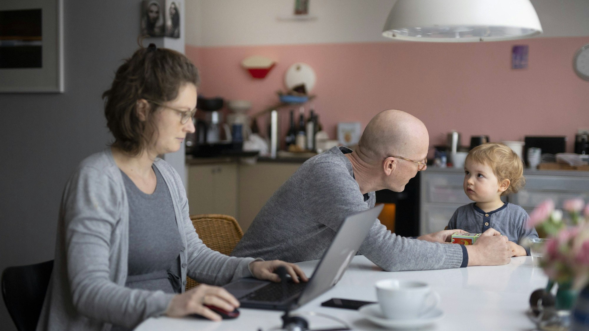 Vater spielt mit Kind, während die Mutter am Laptop arbeitet