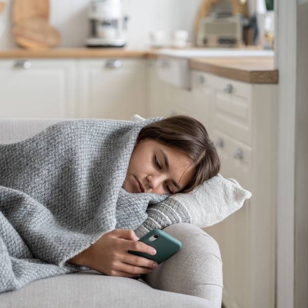 Ein Teenager-Mädchen liegt in eine graue Wolldecke eingewickelt auf dem Sofa und schaut traurig in sein Smartphone