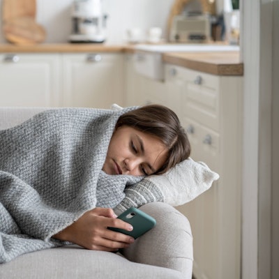 Ein Teenager-Mädchen liegt in eine graue Wolldecke eingewickelt auf dem Sofa und schaut traurig in sein Smartphone