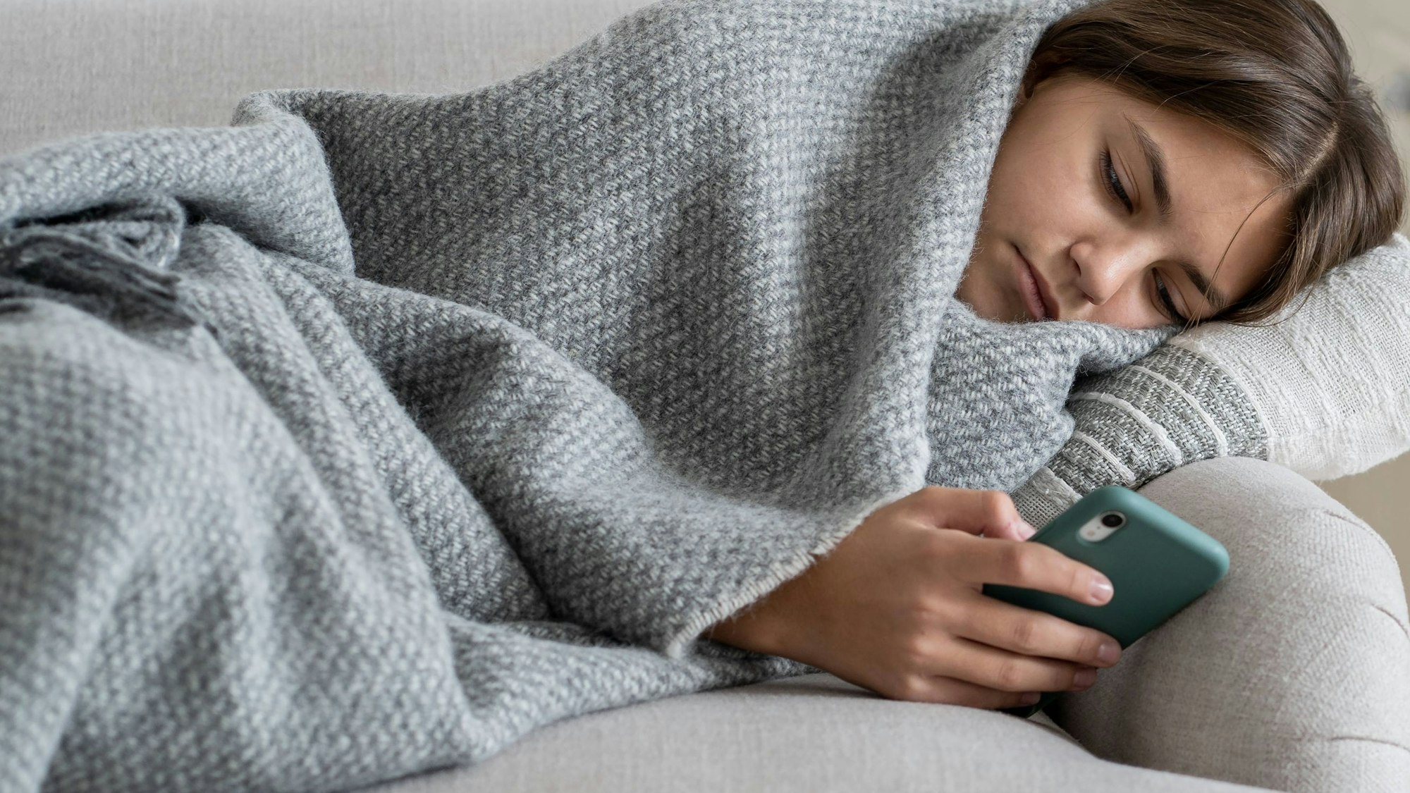 Ein Teenager-Mädchen liegt in eine graue Wolldecke eingewickelt auf dem Sofa und schaut traurig in sein Smartphone