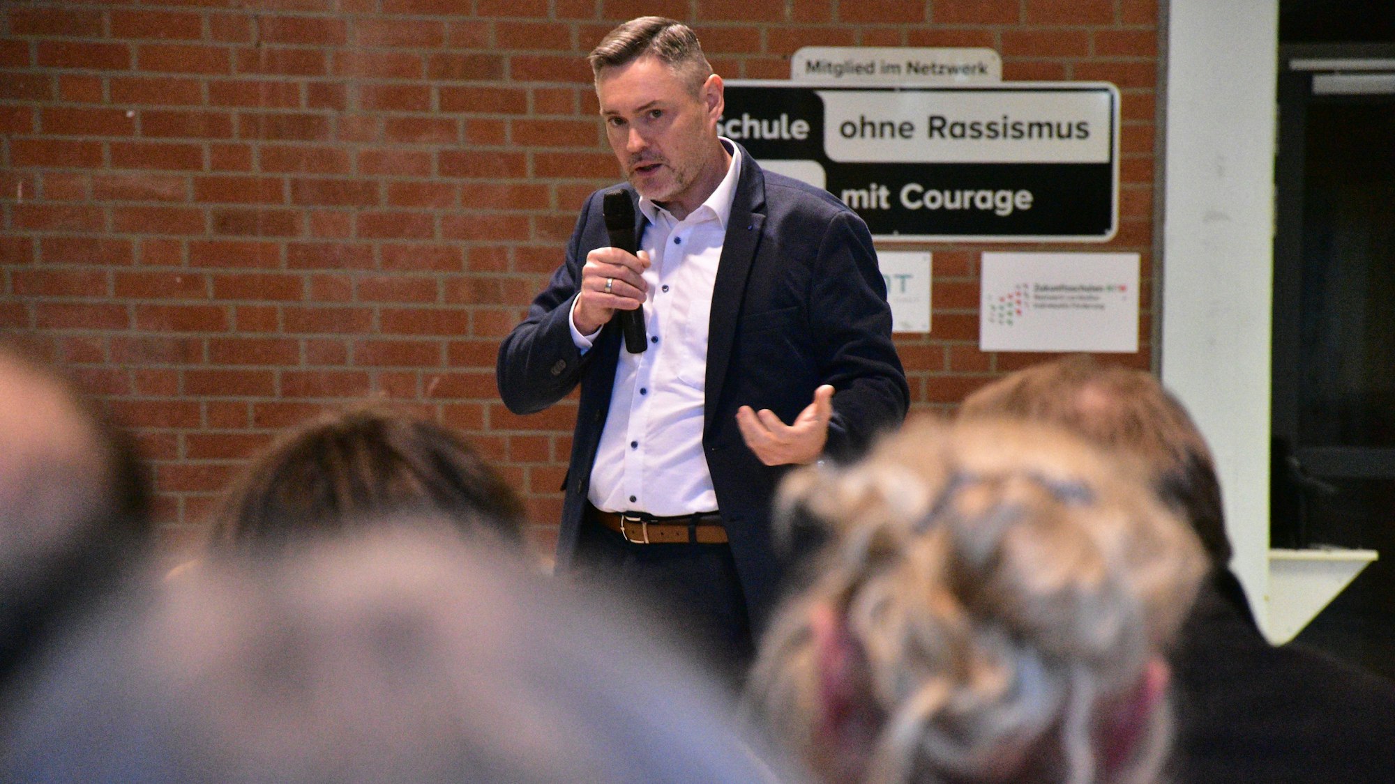 Ein Mann im dunklen Sakko und weißen Hemd spricht vor sitzenden Zuhörern; in der Hand hat er ein Mikrofon.