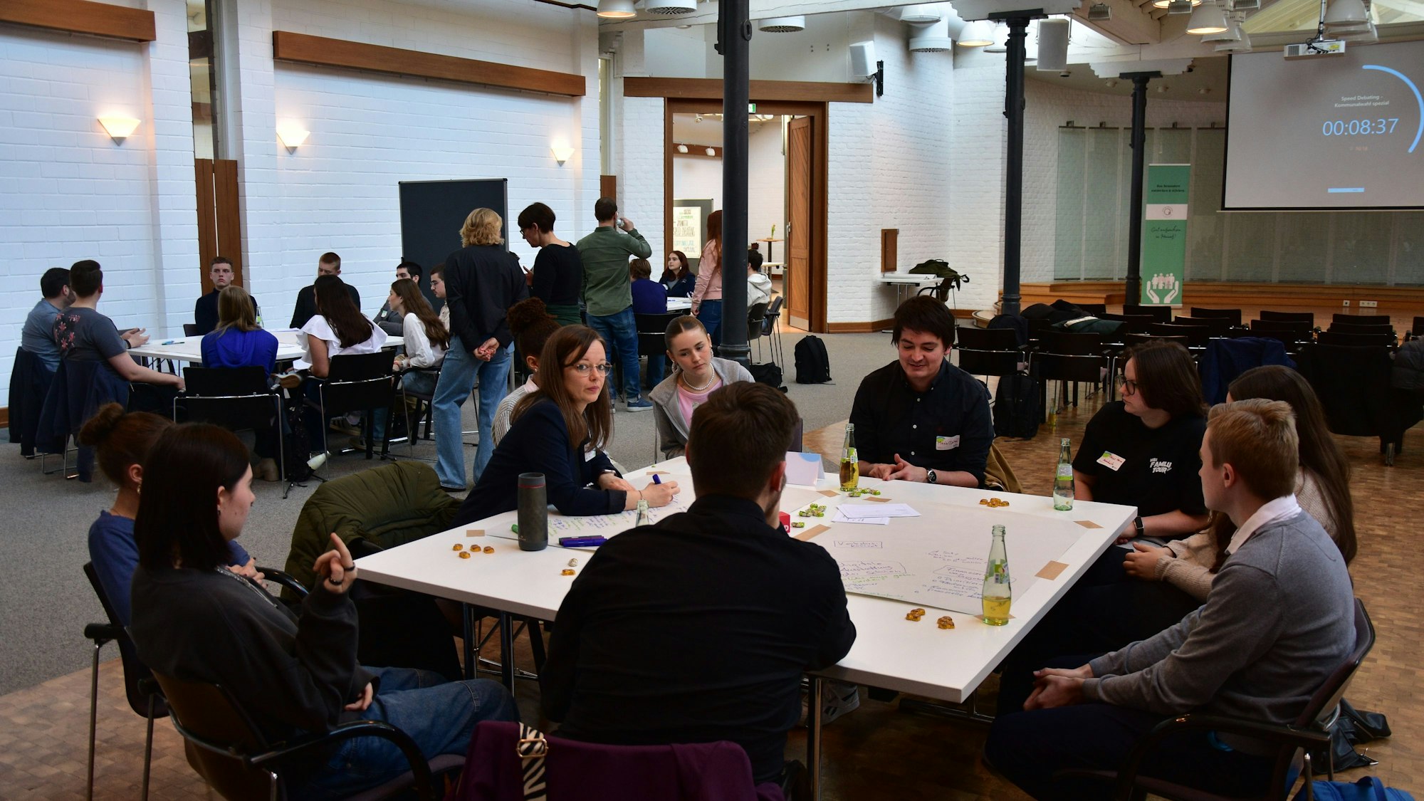 Jugendliche und Erwachsene sitzen an Tischen in einem Veranstaltungssaal