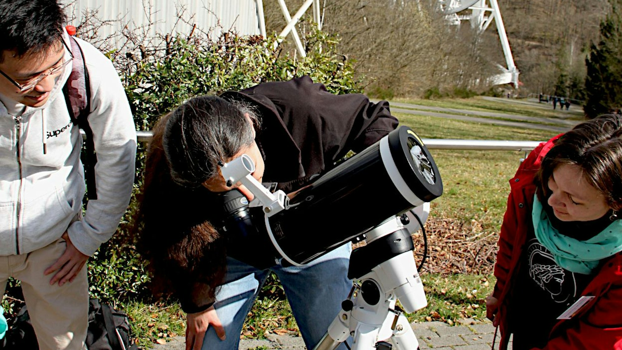 Ein Mann schaut durch ein Teleskop. Ein weiterer Mann und eine Frau stehen rechts und links von ihm und schauen dabei zu.