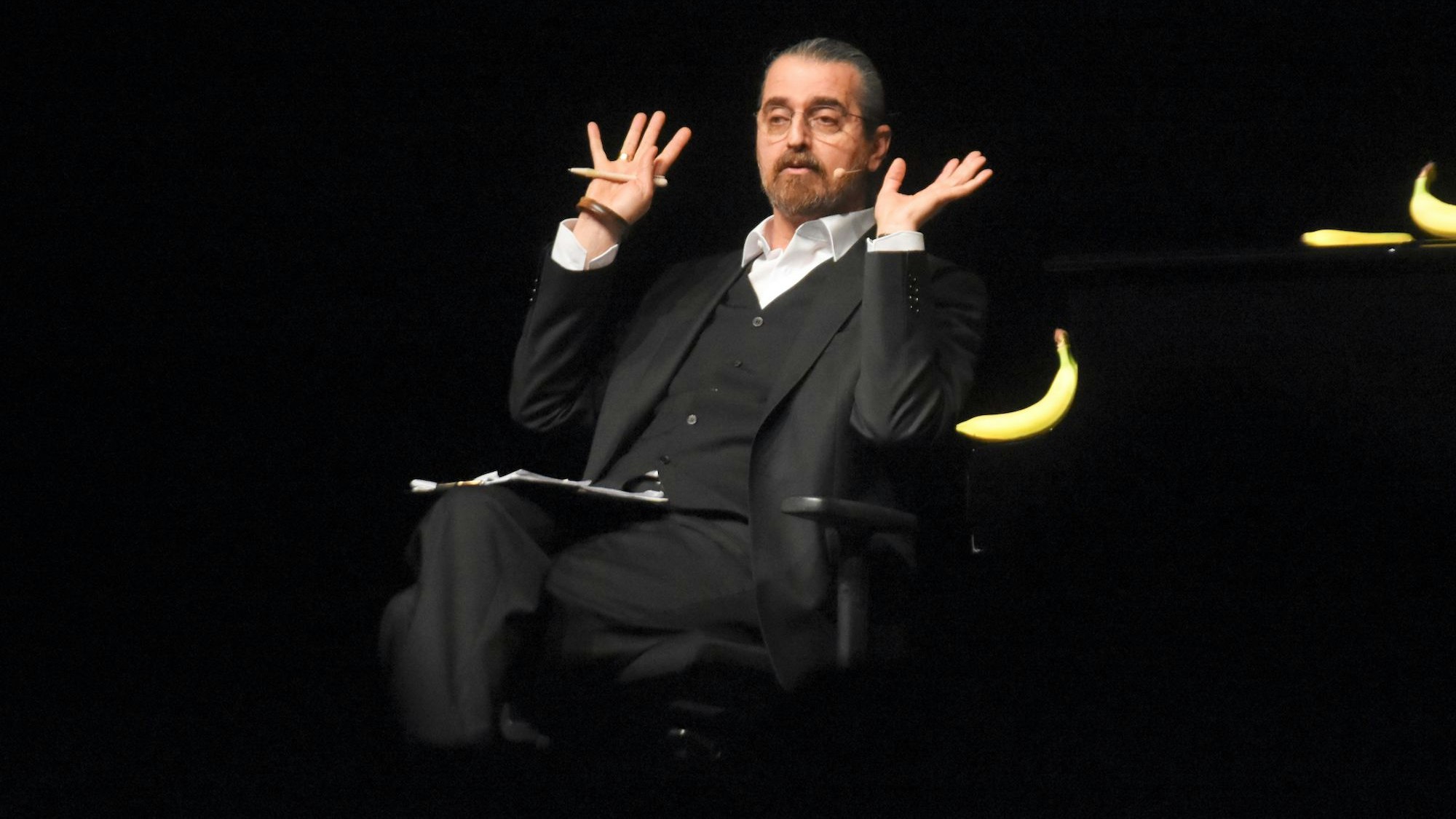 Ein Mann im dunklen dreiteiligen Anzug sitzt auf einem Schreibtischstuhl, er hebt entschuldigend seine Hände.