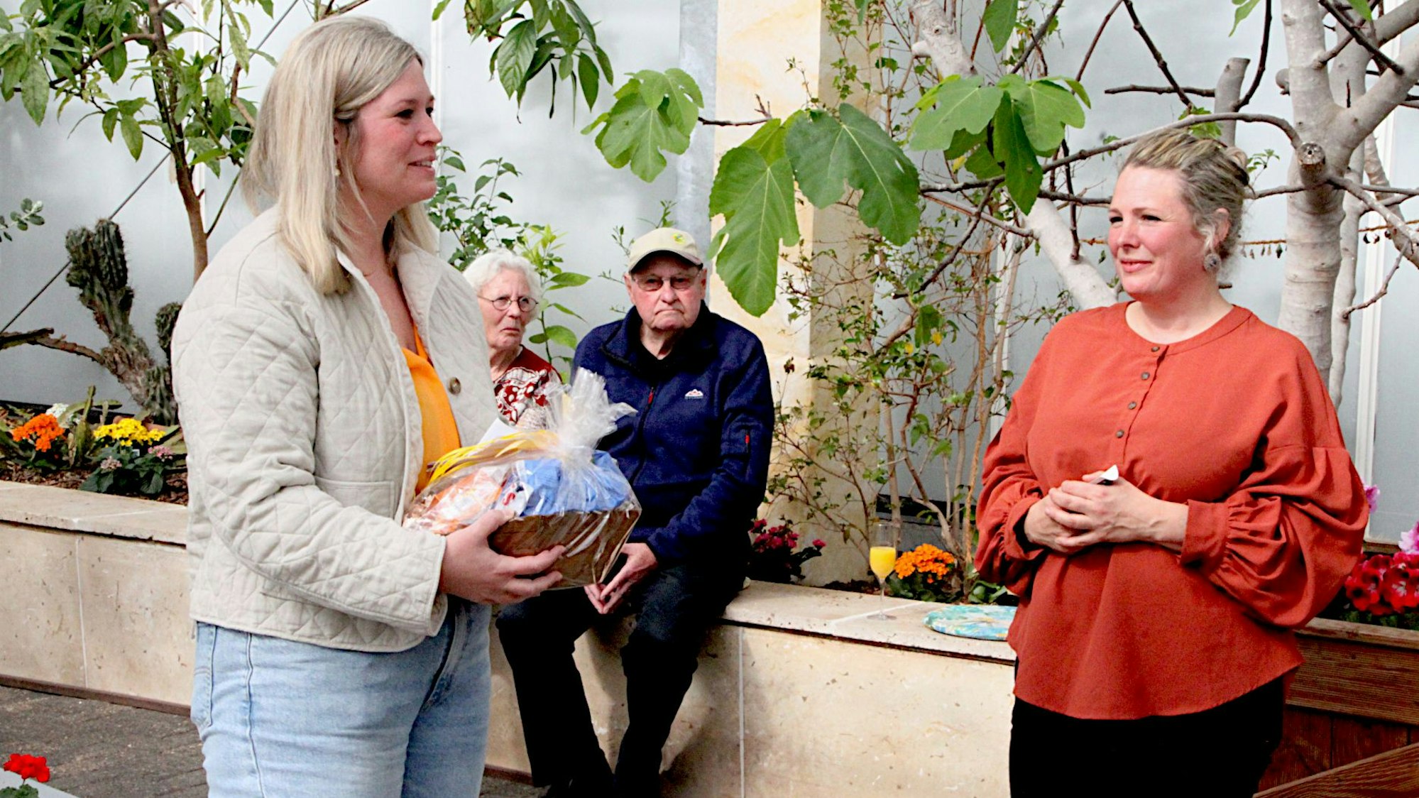 Zwei Frauen schauen sich an, die eine überreicht der anderen ein Geschenk. Im Hintergrund schauen zwei ältere Menschen zu.
