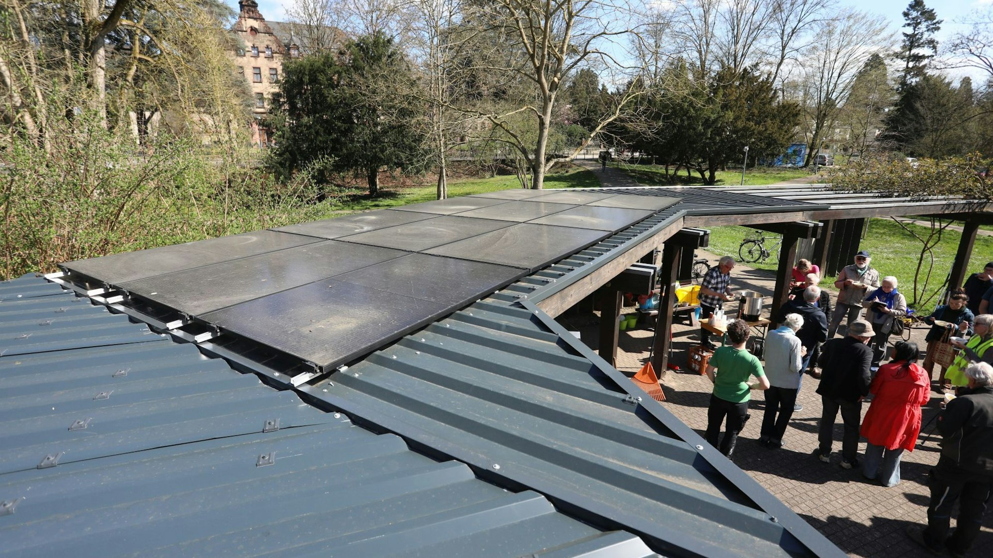 Das mit Metall bedeckte Dach einer Pergola. Darauf sind Module einer Phovoltaikanlage montiert. Am Boden steht eine Gruppe von Menschen.