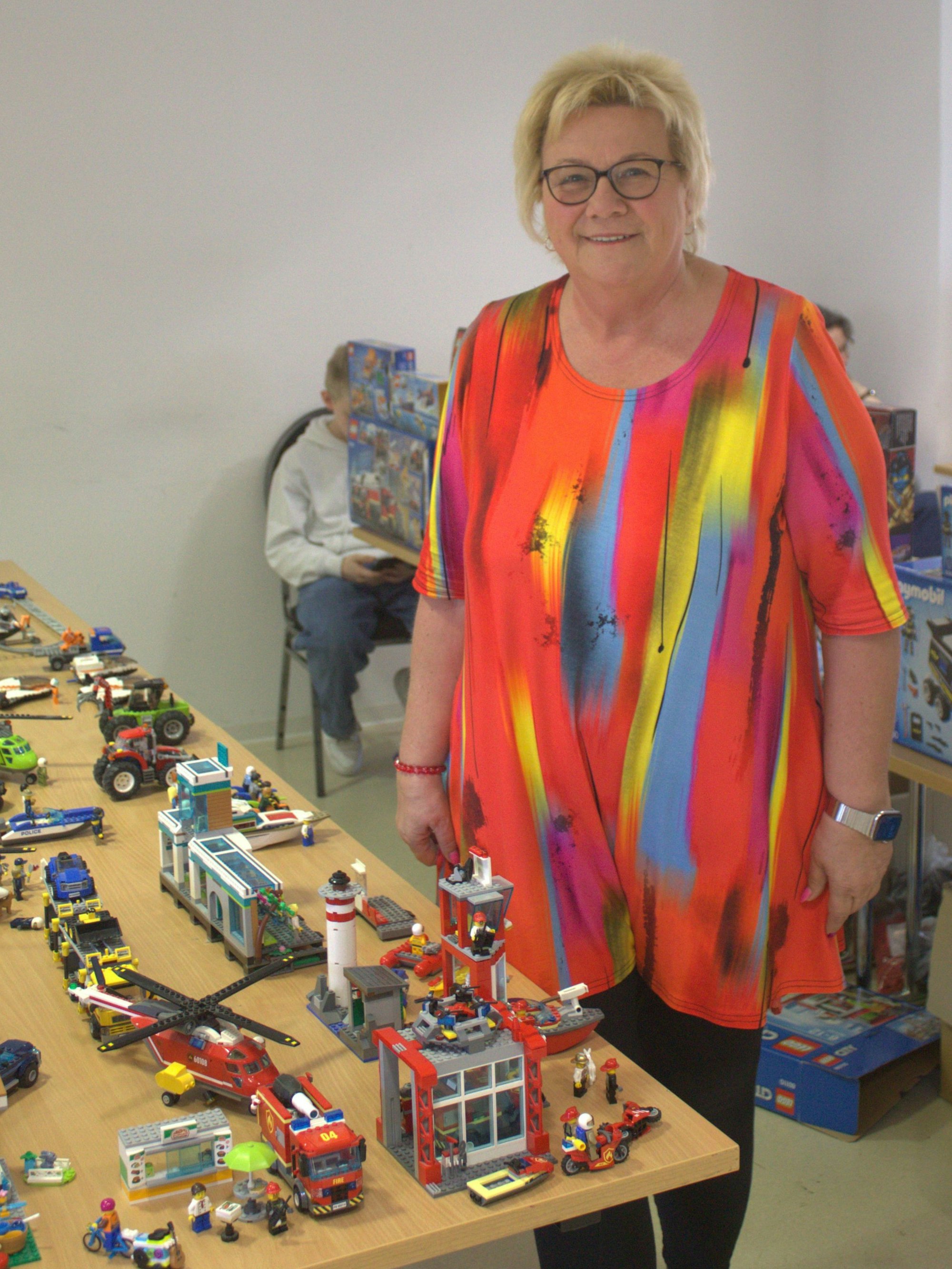 Eine blonde Frau in einer bunten Bluse steht neben einem Tisch mit Lego-Modellen.