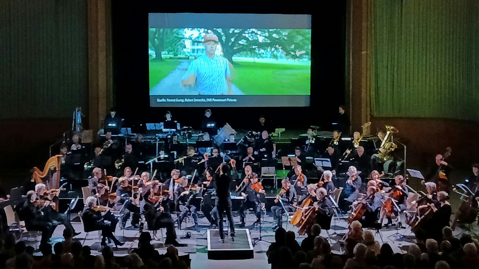 Über einem großen Orchester ist auf der Leinwand ein Ausschnitt aus dem Film „Forrest Gump“ zu sehen.