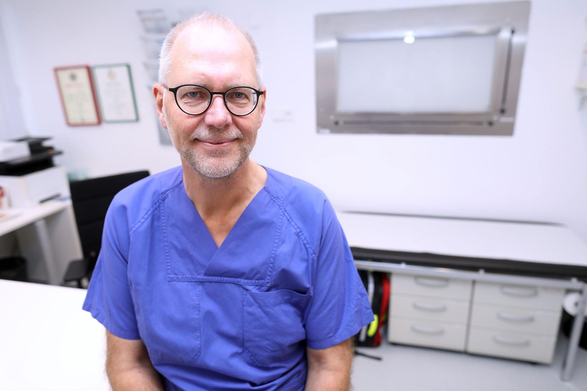 Professor Arno Dormann ist in einem Behandlungsraum zu sehen in blauer Klinikkleidung. Er lächelt in die Kamera.