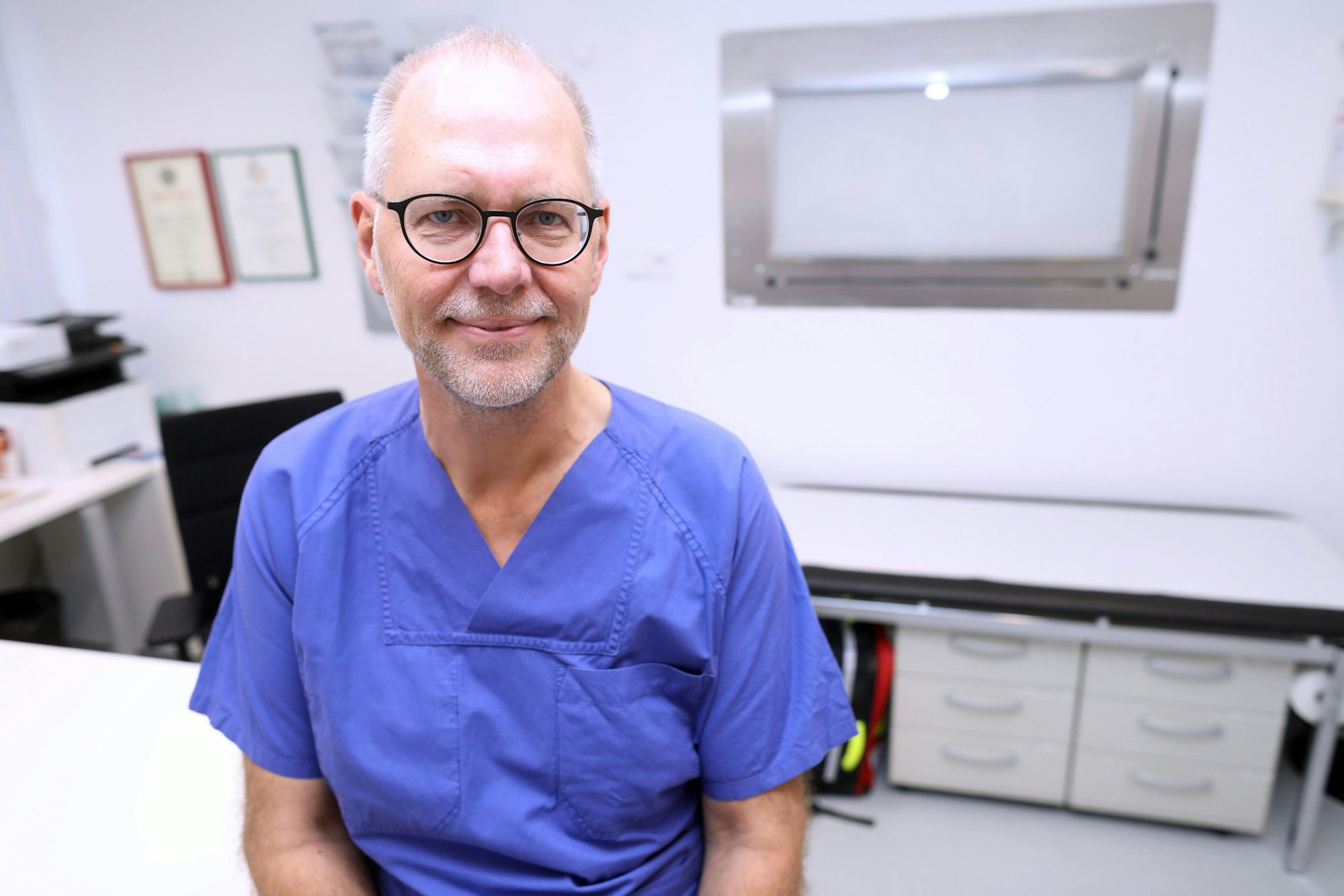 Professor Arno Dormann ist in einem Behandlungsraum zu sehen in blauer Klinikkleidung. Er lächelt in die Kamera.