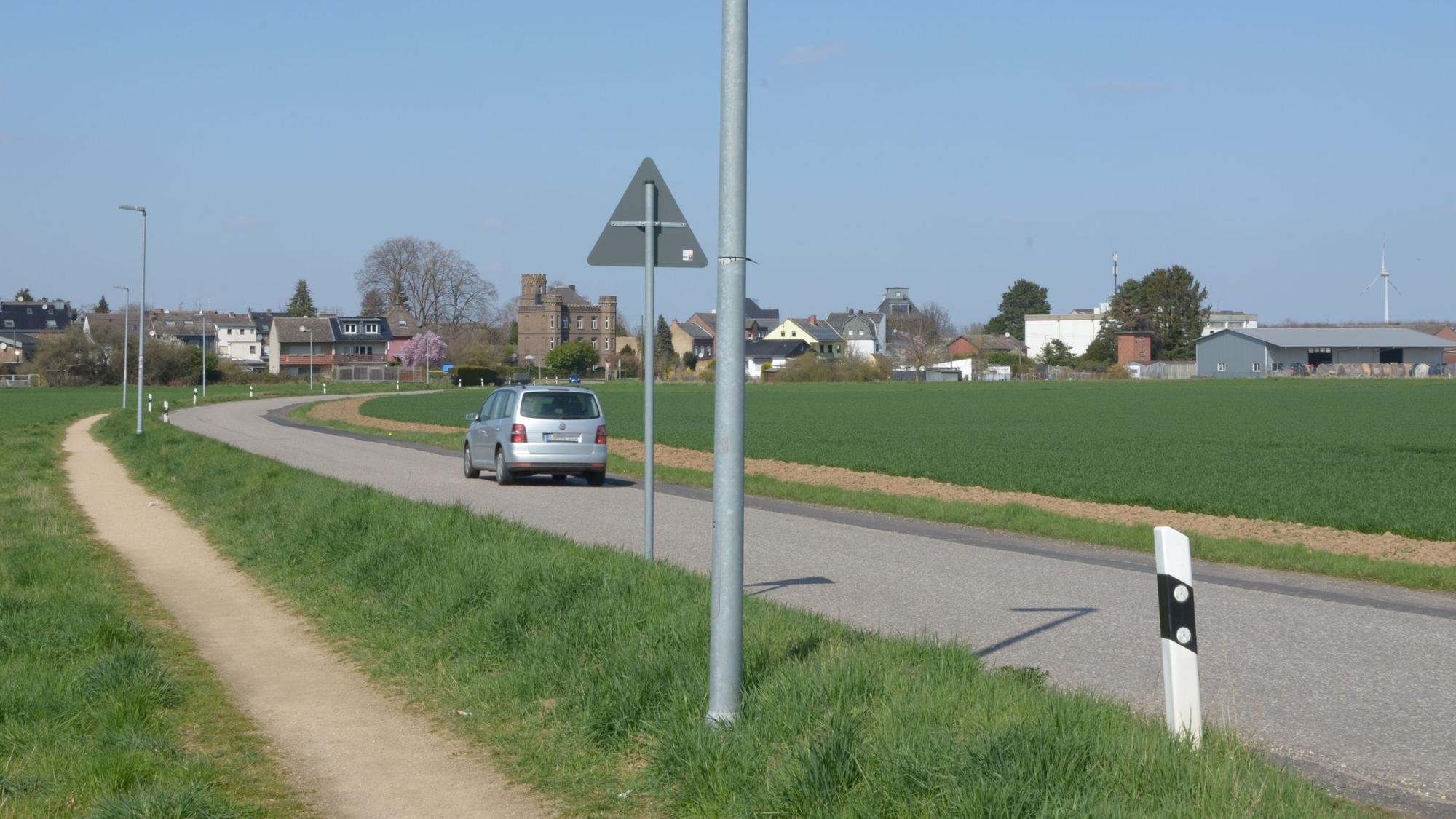 Das Bild zeigt die Heinrich-Ruster-Straße, auf der ein Auto fährt. Neben der Fahrbahn verläuft ein Schotterweg für Fußgänger.
