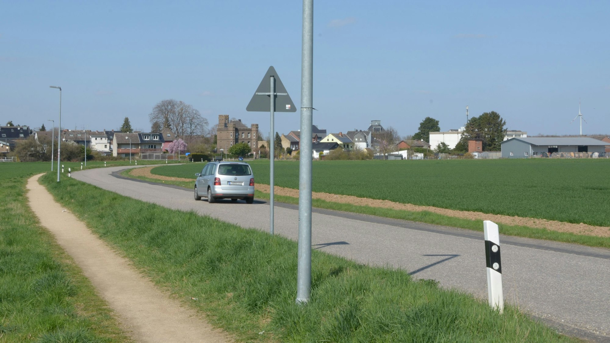 Das Bild zeigt die Heinrich-Ruster-Straße, auf der ein Auto fährt. Neben der Fahrbahn verläuft ein Schotterweg für Fußgänger.