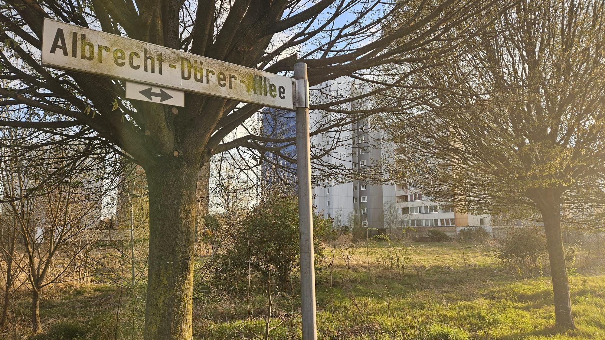 Zu sehen ist ein Straßenschild und dahinter eine brachliegende Wiese sowie Hochhäuser.