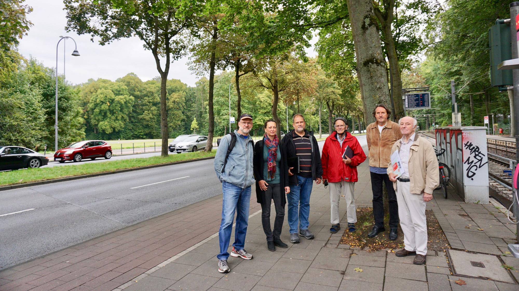 Fünf Männer und eine Frau stehen auf dem Bürgersteig an einer vierspurigen Straße, rechts von ihnen ist eine Straßenbahnhaltestelle zu sehen.