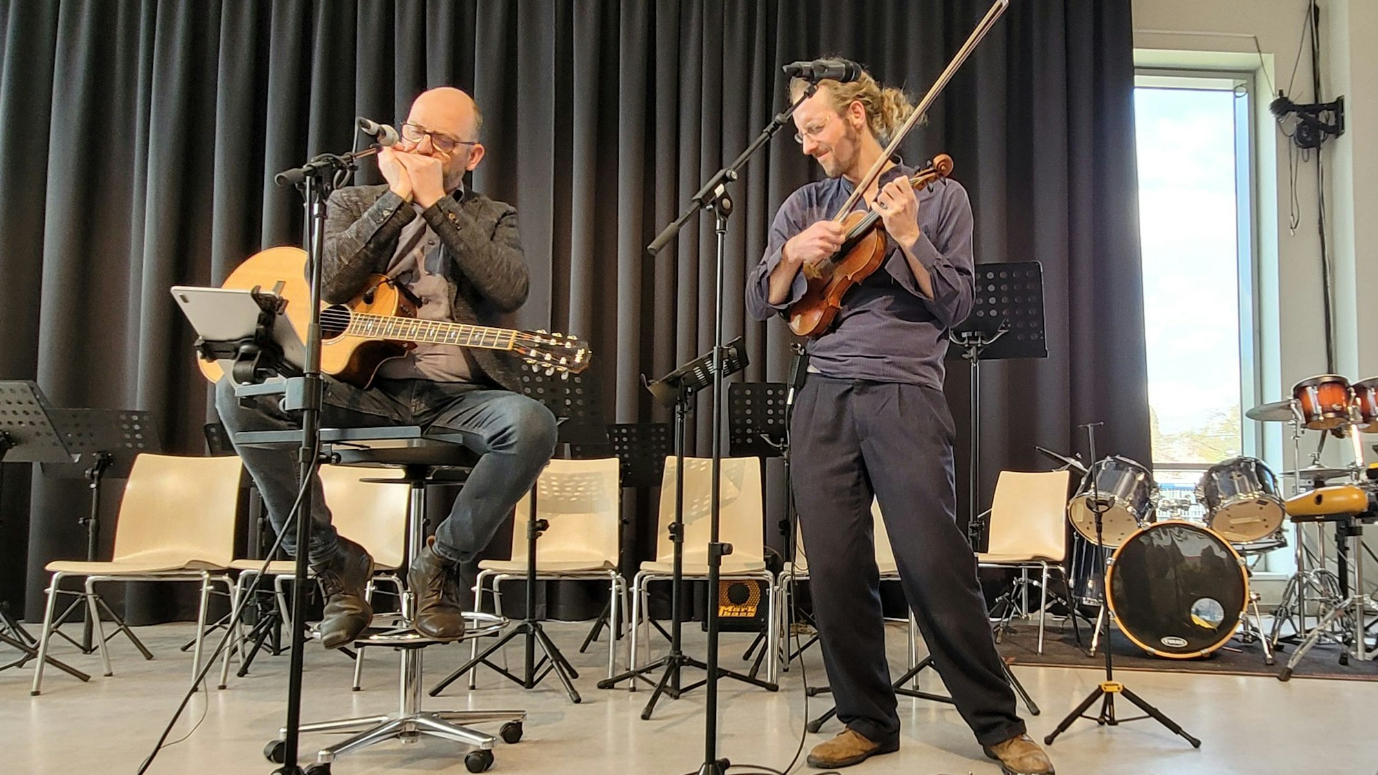 Ein Mann mit Gitarre und Mundharmonika, der zweite, er steht rechts, hält eine Geige in der Hand.
