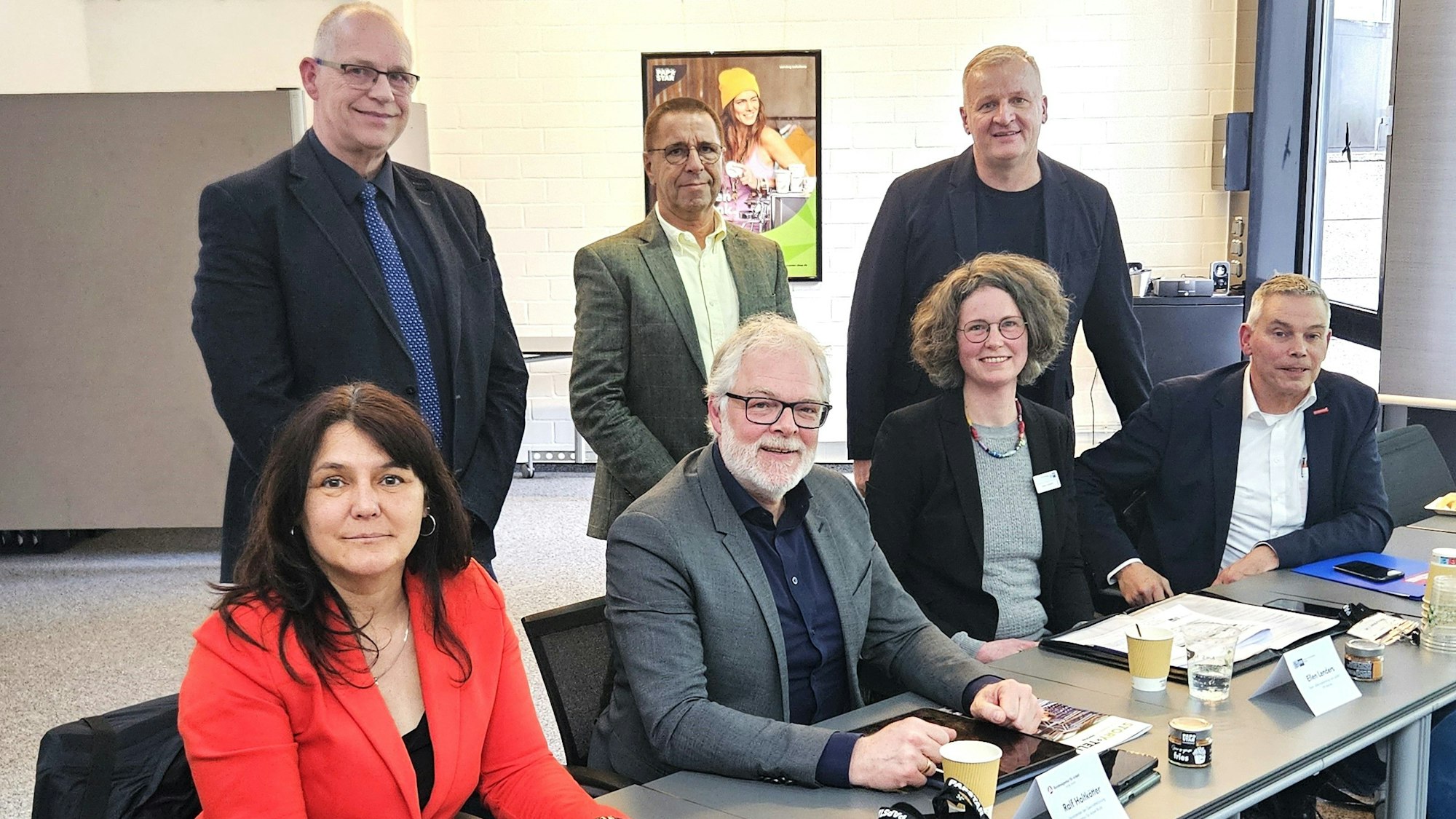 Fünf Männer und zwei Frauen in Anzügen sitzen und stehen an einem Tisch.