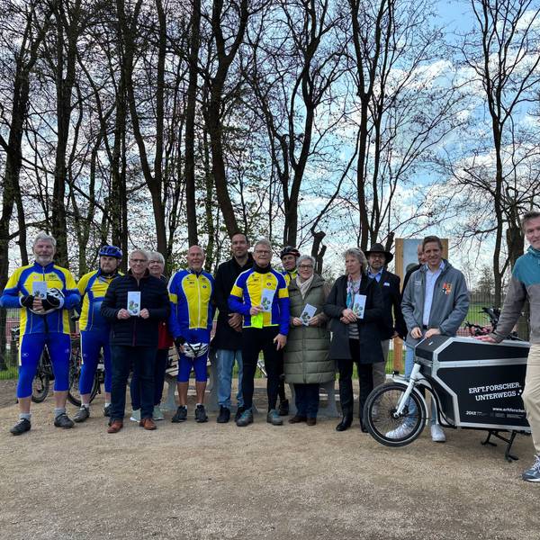 Eine Gruppe, teils in Fahrradanzügen, posiert im Freien vor einem Schild.