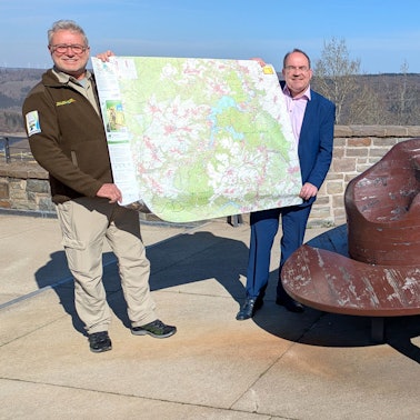 Michael Lammertz und Ralf Nolten (v.l.) präsentieren die neue Nationalpark-Wanderkarte; Jasmin Daus sitzt auf einer Holzbank, die einem Rangerhut nachempfunden ist