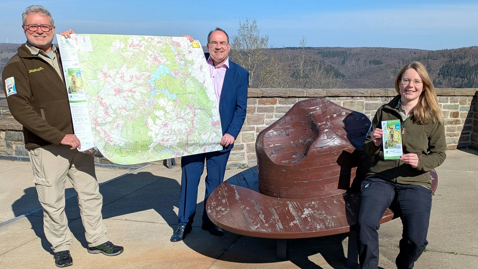 Michael Lammertz und Ralf Nolten (v.l.) präsentieren die neue Nationalpark-Wanderkarte; Jasmin Daus sitzt auf einer Holzbank, die einem Rangerhut nachempfunden ist