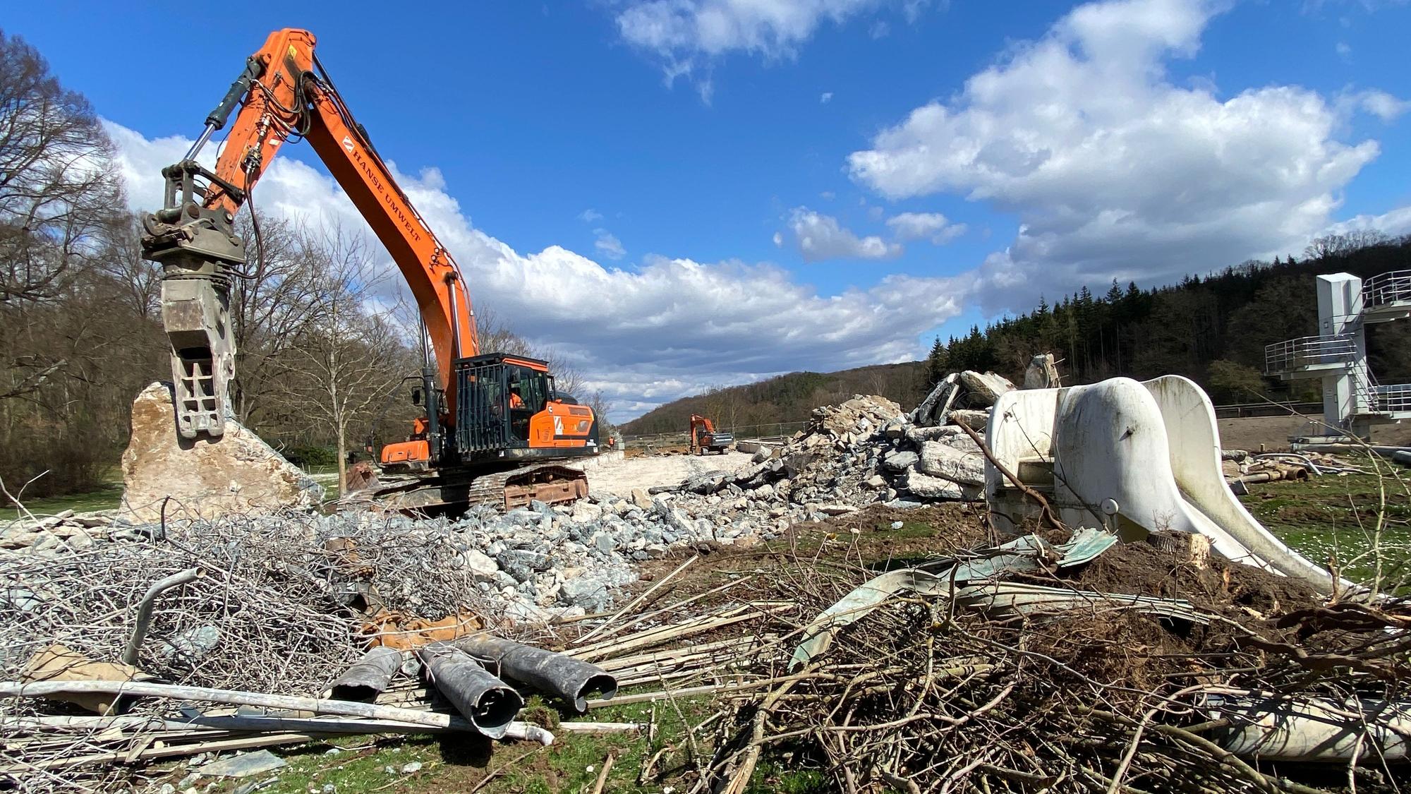 Ein Bagger zerkleinert große Betonstücke. Auf dem Boden liegt Schutt und Metallschrott. Mitten drin steht eine weiße Elefantenrutsche.